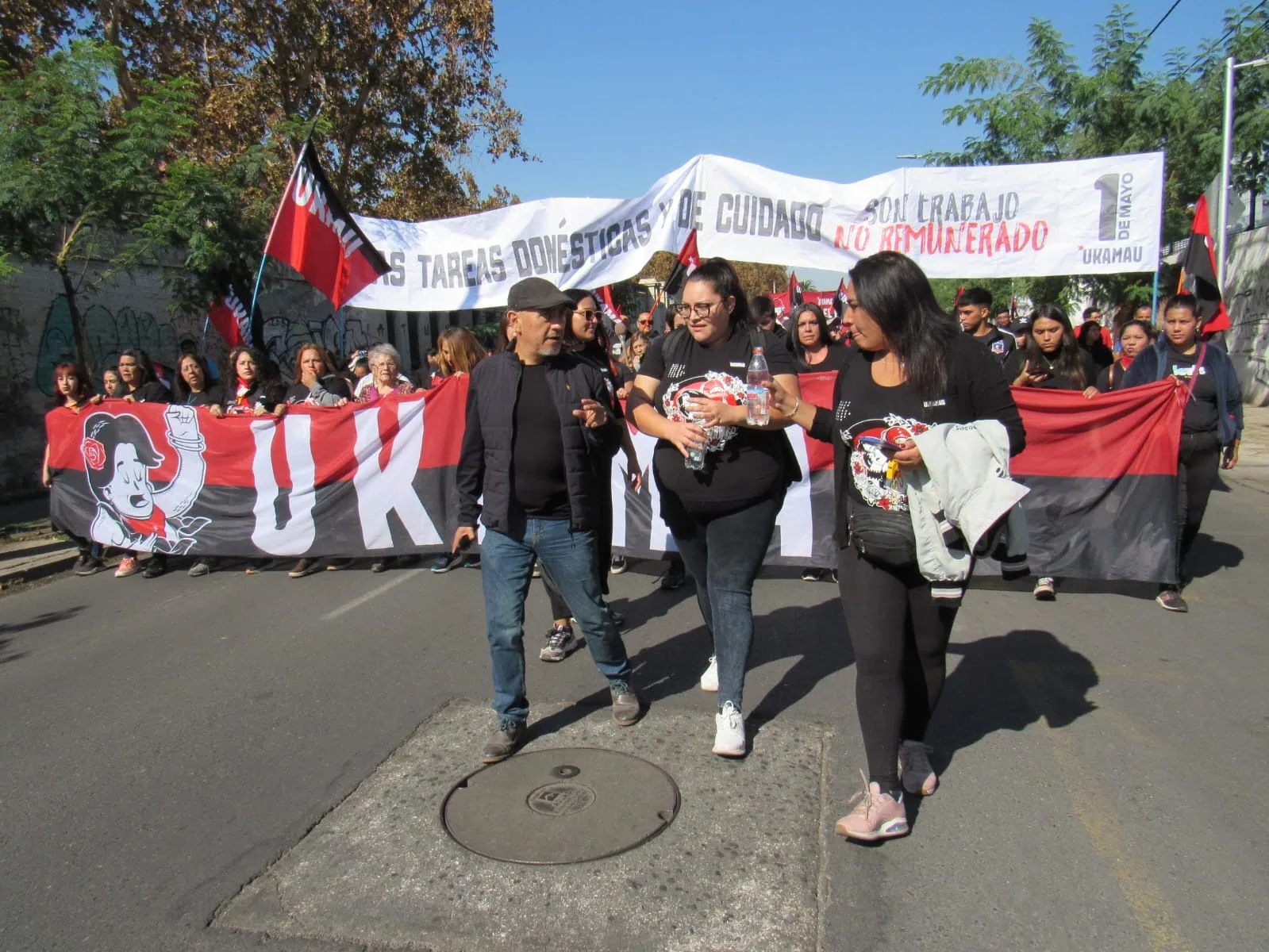 Marcha del Movimiento de Pobladores Ukamau por una calle de Santiago con lienzo rojo y negro y una pancarta sobre tareas domésticas y de cuidado, 1 de mayo