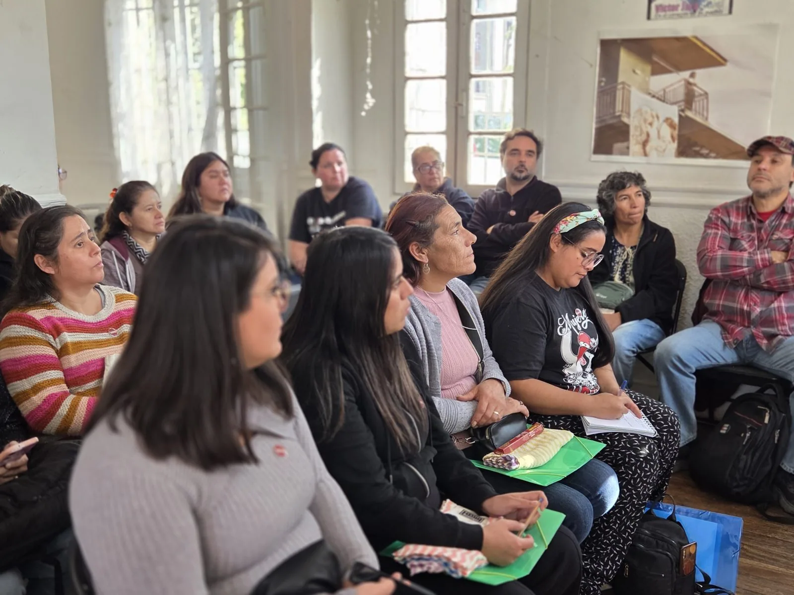 Vecinas y vecinos sentados en una sala durante una sesión de formación, tomando notas sobre carpetas.