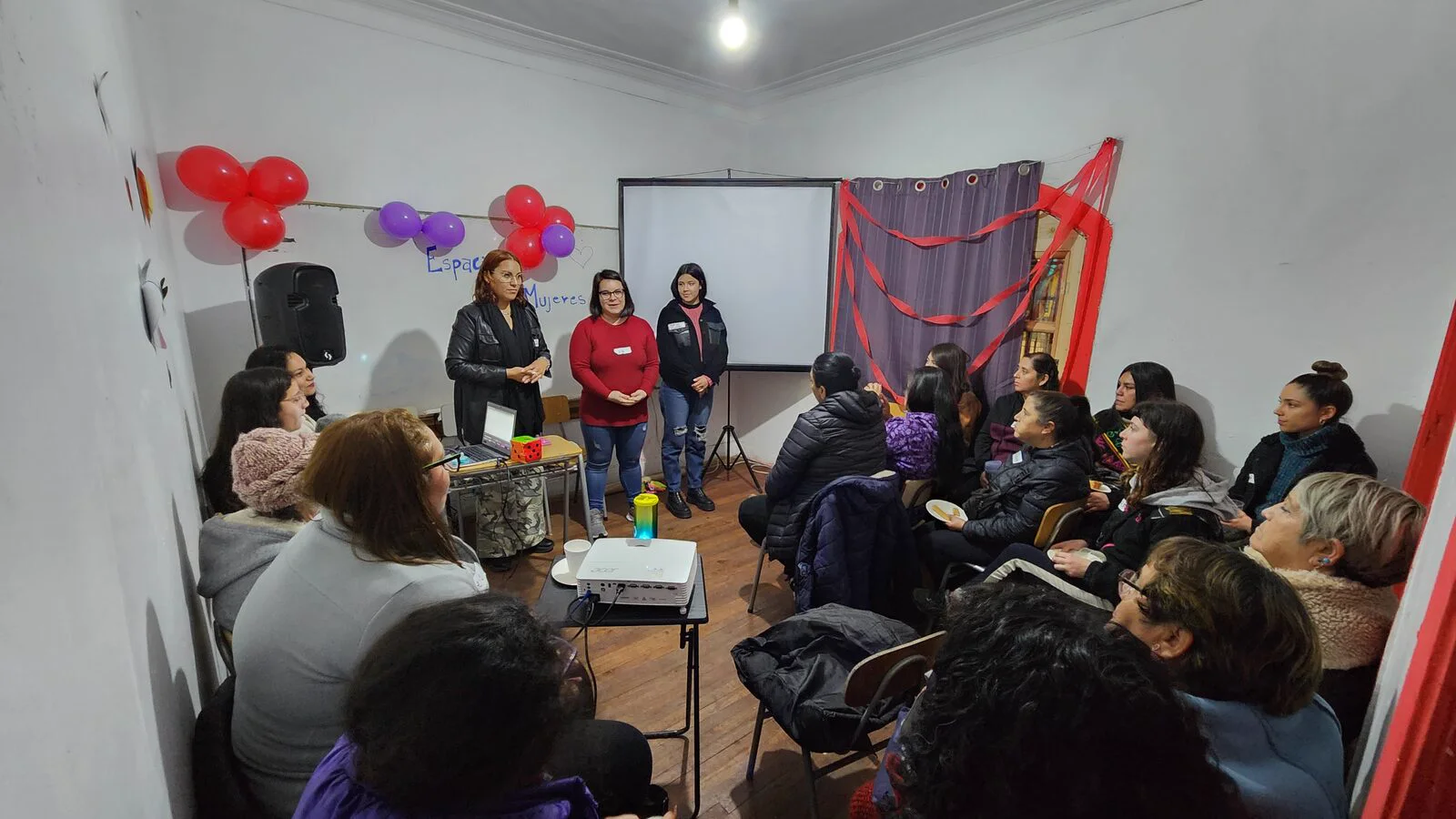 Encuentro de mujeres del comité de vivienda Tercera Etapa: dos monitoras exponen de pie frente a una veintena de mujeres sentadas en una sala decorada con globos.